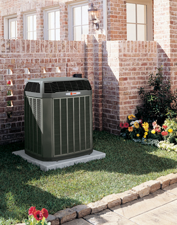 Central air conditioning unit on concrete pad beside brick home, surrounded by green lawn and flower bed.