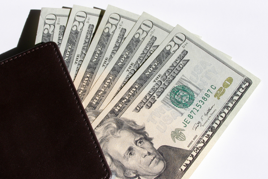 Brown leather wallet partially open with crisp twenty-dollar bills fanned out on well-lit surface.