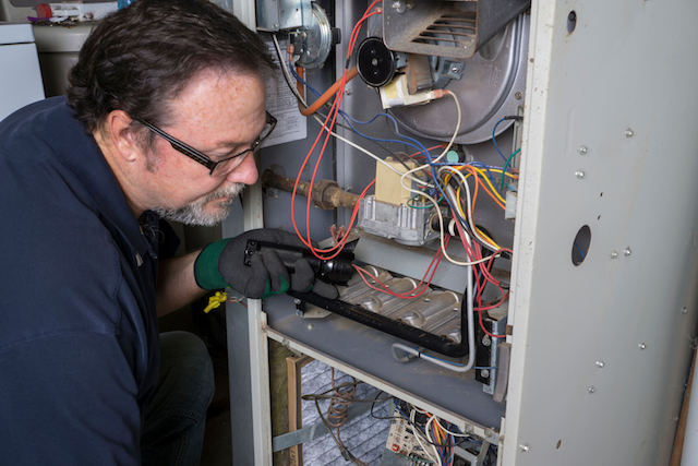 Technician with safety gear using flashlight to inspect internal components of open furnace system.