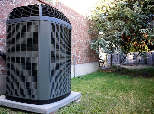 Large outdoor AC unit on concrete pad next to brick building, surrounded by grass, fence, and patio furniture.