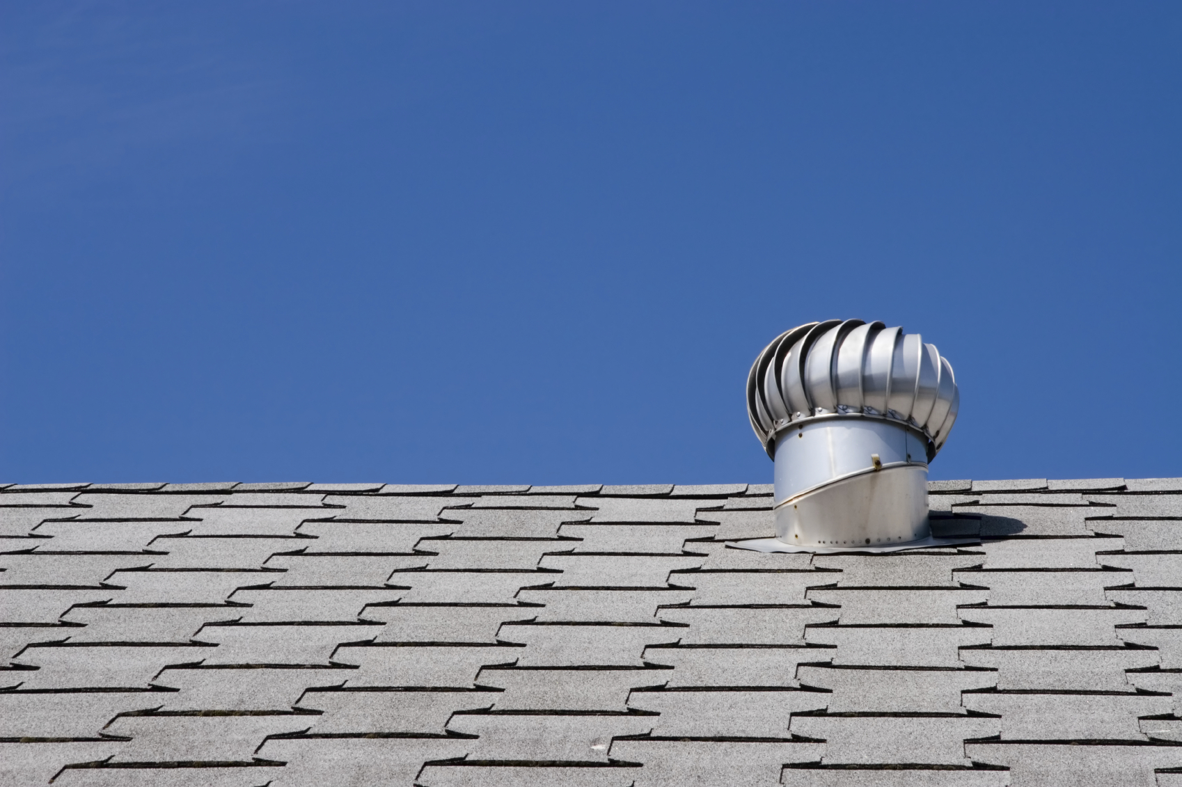 Gray shingle roof with silver turbine vent under clear blue sky, designed for attic ventilation.