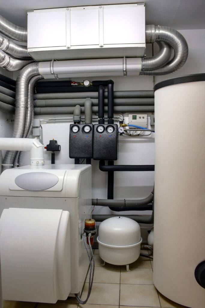 Mechanical room with white boiler, hot water tank, insulated pipes, pressure gauges, and control panels.