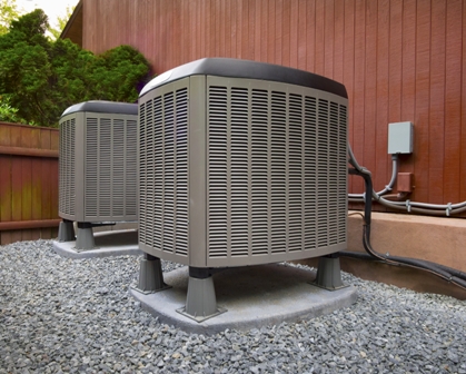 Two outdoor air conditioning units on concrete pads beside wood-paneled building, surrounded by gravel and greenery.