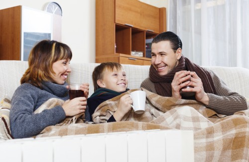 Three people bundled under plaid blanket on couch, sipping hot drinks in warm indoor setting near radiator.