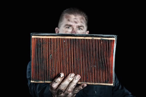 Grease-smudged person holding dirty orange pleated air filter in front of camera against black background.