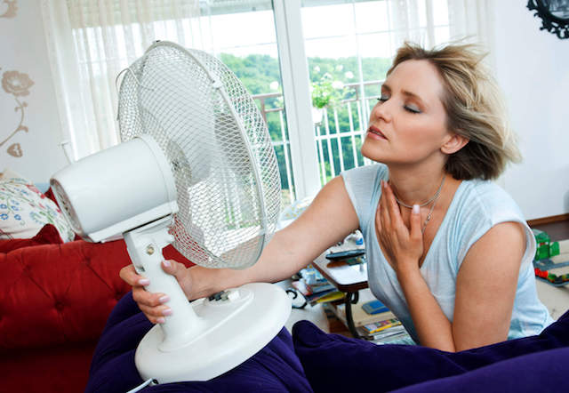 Person sitting indoors with eyes closed, leaning toward electric fan on red couch to cool off.
