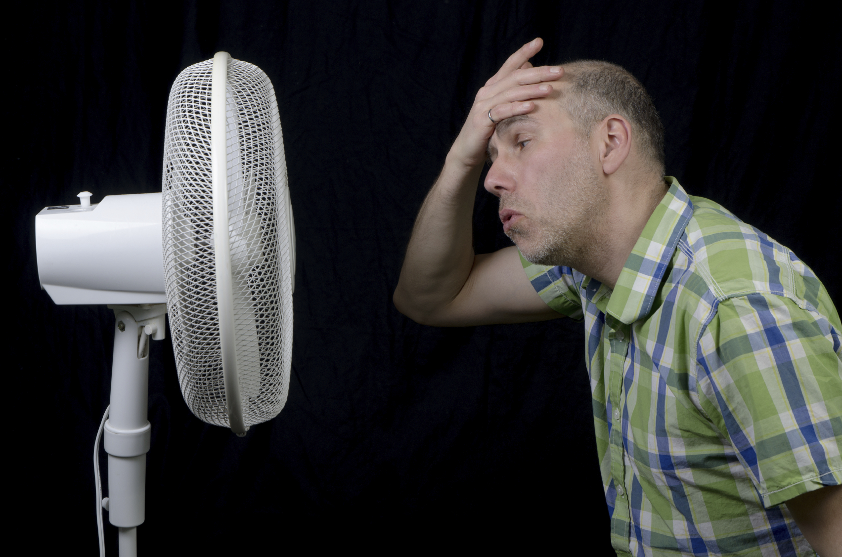 Person in plaid shirt cooling off in front of white oscillating fan, leaning forward with hand on forehead.