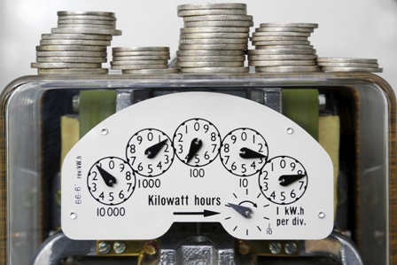 Analog electricity meter with five dials and stacked coins, symbolizing kilowatt-hour consumption and cost.