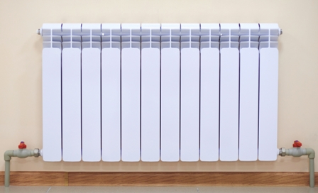 White vertical-panel radiator with dual red-valve pipe connections mounted on beige wall above wood baseboard.