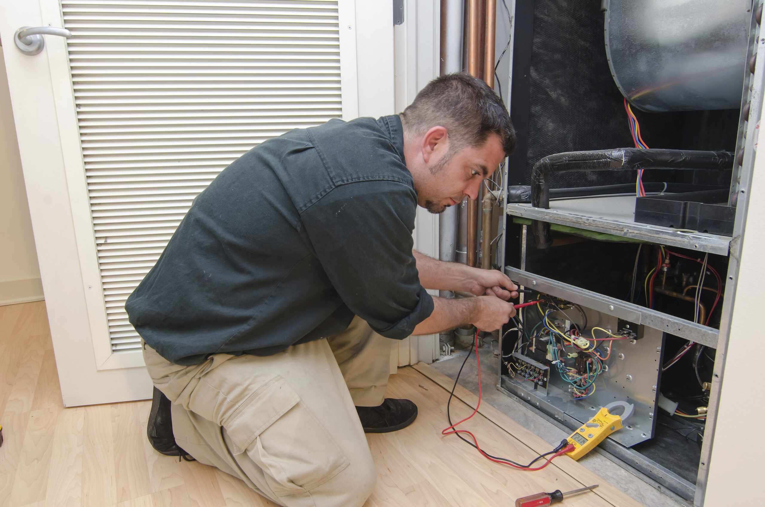Technician kneeling beside open HVAC panel, using multimeter to test wiring and circuit boards.