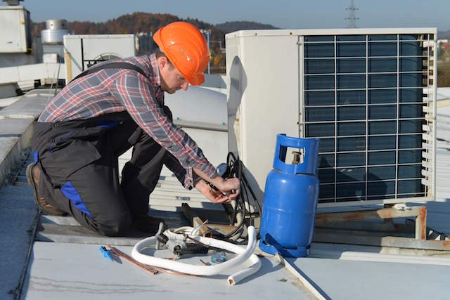 Technician in helmet and overalls servicing rooftop HVAC unit with tools, refrigerant cylinder, and insulated pipes.